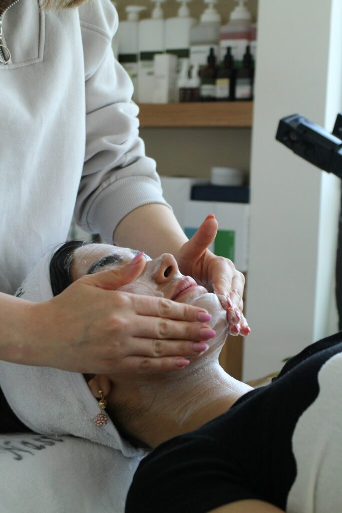 An esthetician gently applying a soothing facial mask to a person with visible delicate skin in a calm spa room.